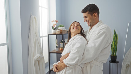 Man and woman in white robes smiling and embracing in a wellness spa room, with bright natural light coming through the windows and cozy interior decor