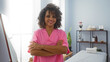 © Krakenimages.com - Woman with curly hair and pink uniform standing with arms crossed in a spa room, indicating wellness and beauty services provided by a smiling professional in an indoor salon setting