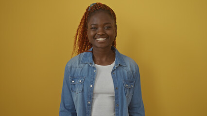 A cheerful young african woman in casual denim attire posing against a vibrant yellow background with a genuine smile.