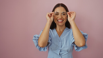 Beautiful young hispanic woman in blue dress adjusting glasses with a bright smile, standing over an isolated pink background wall