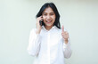© M Alfan Setyawan - portrait of successful confident asian young woman entrepreneur speaking on smartphone and looking at camera giving thumbs up feeling satisfied of service wearing white formal shirt, standing isolated