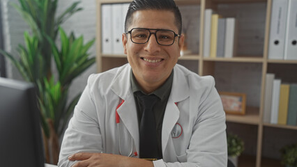 Smiling hispanic man wearing glasses and lab coat seated indoors at clinic workplace.