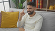 © Krakenimages.com - A smiling young hispanic man with a beard gestures while sitting on a sofa in a cozy living room setting.