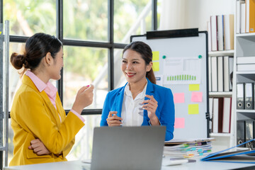 Collaborative Brainstorm: Two young Asian businesswomen engage in a lively discussion at a desk in a modern office, showcasing their teamwork and innovative ideas.  