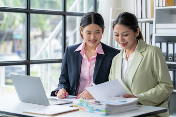 Collaborative Success: Two smiling Asian businesswomen review documents together in a modern office, showcasing teamwork and professional collaboration.