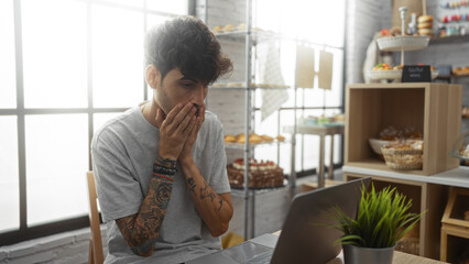 Wall Mural - Young man with a beard and tattoos looking surprised while sitting in a bakery cafe with a laptop on the table