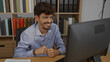 © Krakenimages.com - Young hispanic man with glasses and beard smiling while working on computer in an office environment with shelves of books in the background