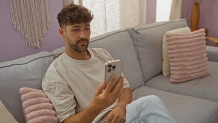 Wall Mural - Young man sitting on a couch at home, looking at his smartphone in a modern living room.