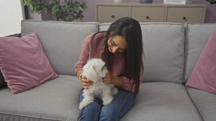 Wall Mural - Hispanic woman affectionately cuddling her white bichon maltese dog on a grey couch indoors