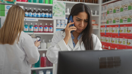 Two women pharmacists in white coats work in a drugstore, one consulting on a phone, the other using a tablet