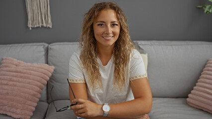 Wall Mural - Smiling young woman holding glasses with arms crossed in a comfortable living room setting with cushions on a sofa.