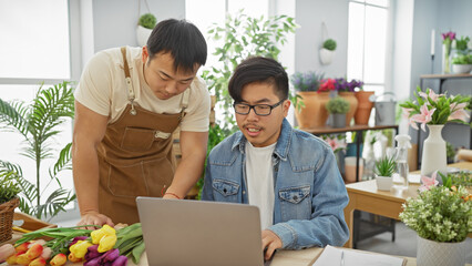 Two men collaborate in a flower shop, discussing over a laptop amid fresh blossoms and plants.