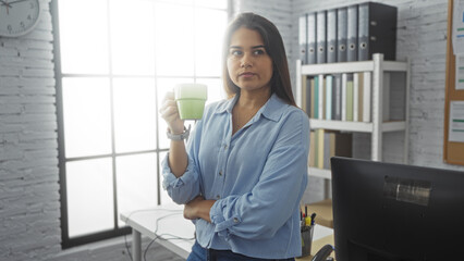 Woman drinking coffee in an office, standing by a desk with a computer, looking thoughtful, wearing a blue shirt and surrounded by shelves and files.