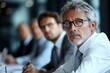 © Odin AI - A senior business leader with gray hair attentively listens during a corporate meeting, wearing glasses and a tie, focused on the discussion at hand.