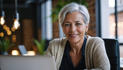 A confident senior woman with gray hair smiling warmly, working on a laptop in a modern and cozy office setting with ambient lighting.

