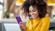 © ChimE - A cheerful woman with curly hair in a yellow shirt focuses on her purple smartphone while balancing work at a cafe.