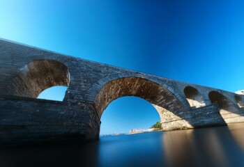 Aufkleber - A stone arch bridge spans a body of water, with a clear blue sky above.