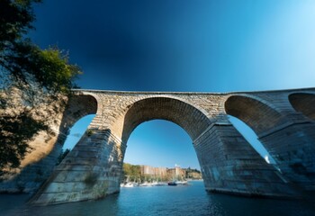 Sticker - A stone arch bridge spans a body of water, with boats and a cityscape in the background, all under a bright blue sky.