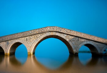Aufkleber - A stone bridge arches over a still body of water with a bright blue sky above.