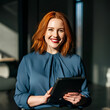 © Paintbrush - A smiling woman with red hair, holding a tablet, standing in a bright office environment.