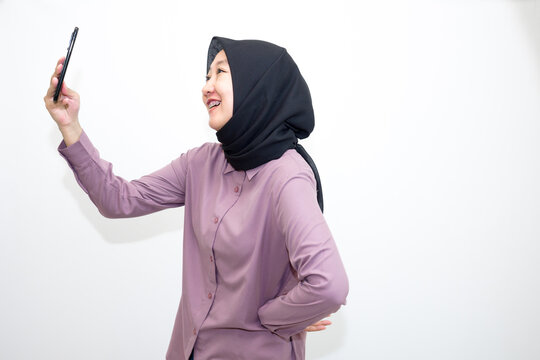 Portrait of emotional Asian, Indonesian woman wearing purple shirt and hijab headscarf laughing and holding handphone. Isolated on white background