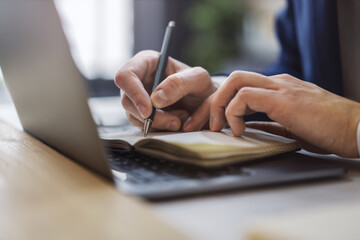 Poster - Detailed view of a man's hand jotting down notes in a notepad on a modern laptop, with a blurred background