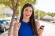 © luismolinero - Young pretty Brazilian woman using mobile phone at outdoors pointing up a great idea