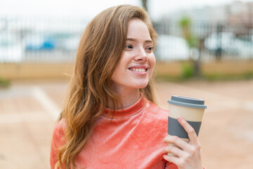 Wall Mural - Young redhead woman at outdoors holding a take away coffee with happy expression