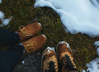 Brown men and women boots standing on wet ground with patches of grass and snow. A winter hiking and travel concept, showcasing outdoor adventure in cold weather conditions