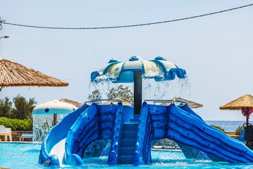 Blue water slide in outdoor pool area with straw umbrellas at beach resort on sunny day.