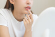 © KMPZZZ - Lip care concept, worried asian young woman finger touching lips in front of mirror after applying lipstick balm on dry mouth from natural beauty product, skincare routine, makeup and cosmetics.