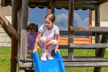 Two young girls playing on a playground slide outdoors, sunny summer holidays day, carefree enjoying childhood fun, outdoor activity, family time, friendship, and healthy lifestyle in a rural setting