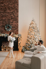 Wall Mural - Young woman hugs her beloved husband giving him a gift on this special day. Concept of gifts for birthdays, Christmas, Valentine's Day and Men's Day. Modern living room interior. . Part of a series