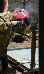 welder in red protective mask welds metal pipe to iron leg of inverted table, welding of iron parts in metal furniture manufacturing process