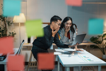 Businessman and businesswoman are working late in the office, collaborating on a project using a laptop and sticky notes