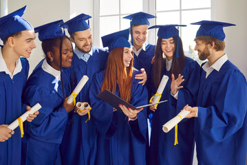 Portrait of a group of smiling happy multiracial international graduates students talking and having fun in a university graduate gown and holding diploma indoors. Education and graduation concept.