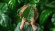 © Maximages  - In a playful interaction with nature, a child hides their face behind a leaf amidst lush green foliage, evoking a sense of innocence and connection to nature.