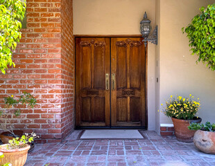 front door of a American house: wooden double doors with intricate carvings and brass doorknobs; brick wall, stucco, rustic antique lantern, welcome mat, potted flowers and plants and trees