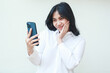 © M Alfan Setyawan - grateful shy asian business woman feeling embarrassed taking selfie and video call on smartphone hold on hands wearing white formal suit shirt standing isolated, online lifestyle concept