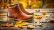 ©  VladaToday - Brown men's boots placed on a wooden terrace with yellow autumn leaves scattered around.