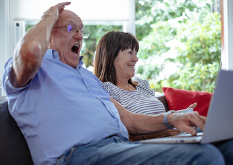 Elderly man sitting on the sofa with his wife while looking surprised at the laptop, two elderly pensioners in their house with a large window on the garden