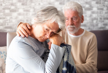 Senior man comforting his depressed sad wife, unhappy elderly woman sitting on sofa at home needed support and care. Two pensioners express their feelings and closeness