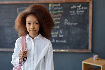 Waist up portrait of cute African American girl with puffy curly hair looking at camera holding backpack and posing against blackboard copy space