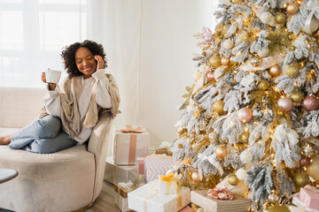 Merry Christmas. African American woman with cup of hot drink coffee tea sitting near Christmas tree at home. Girl in living room with Christmas tree. Christmas hygge holidays at home