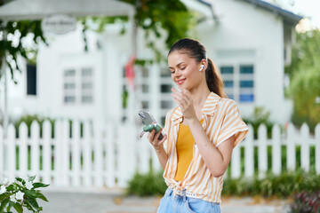 Young woman enjoying music outdoors, smiling while using smartphone in a sunny environment, sporting casual attire with headphones, vibrant green foliage in the background