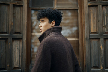 Fashionable young man with curly hair wearing a brown coat, standing in front of an old wooden door and looking to the side