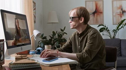 Man with disability making video call with woman while sitting in front of computer reading braille book at desk in living room