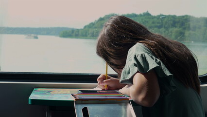 Wall Mural - Young girl drawing with a pencil on a table while traveling by train, looking focused on her work with scenic landscape visible through the window in the background