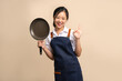 © PBXStudio - Asian woman wearing apron and holding a frying pan. She makes an ok sign on beige background.