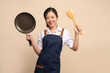 © PBXStudio - Portrait of beautiful Asian woman in apron with black pan and spatula on beige background.